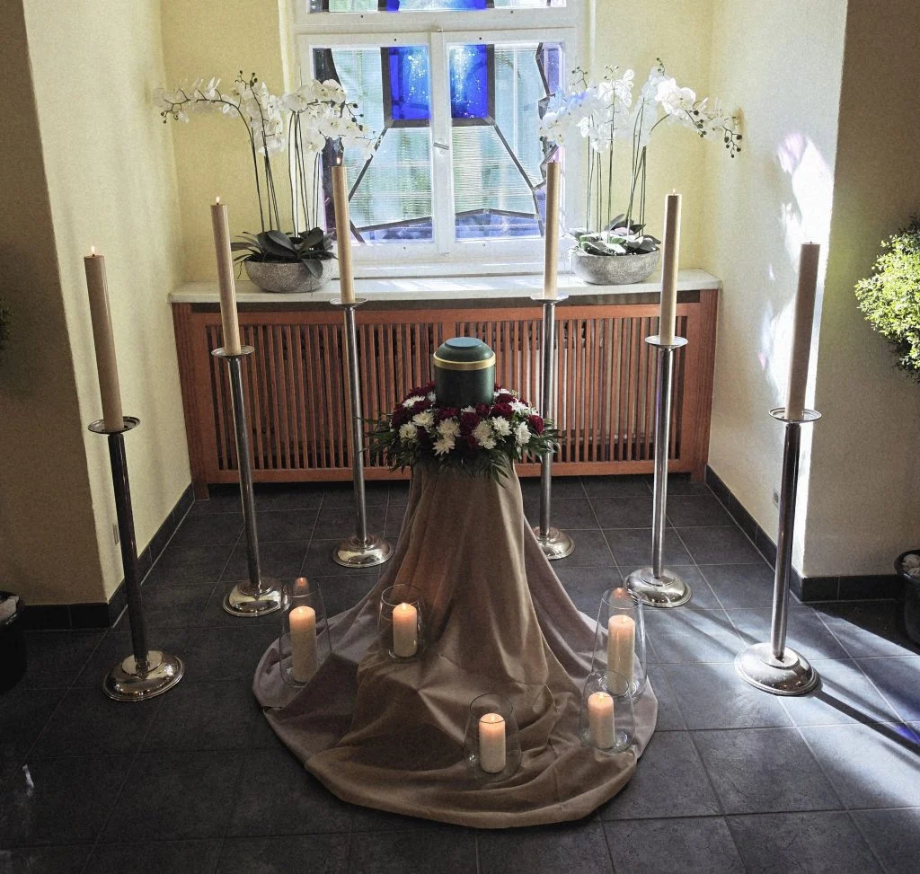 Urne auf einem Podest mit Kerzen, Orchideen und Blumenschmuck in einer Trauerhalle