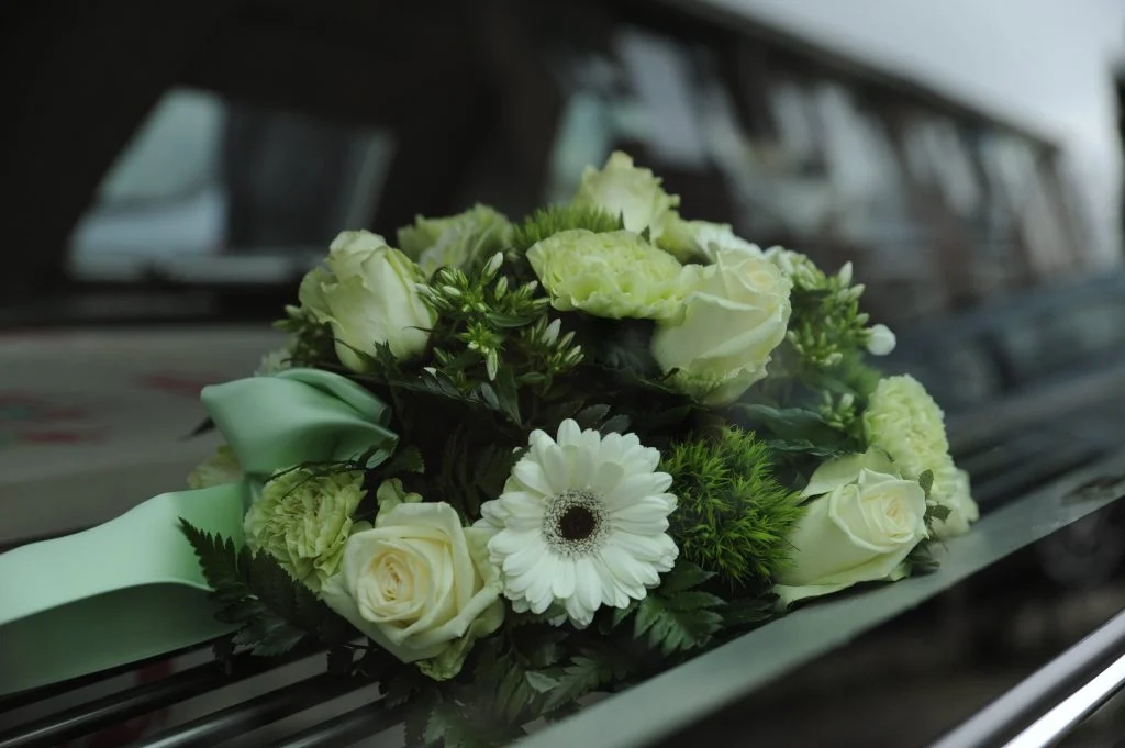 Weißes Blumengebinde mit grüner Schleife auf einem Bestattungswagen